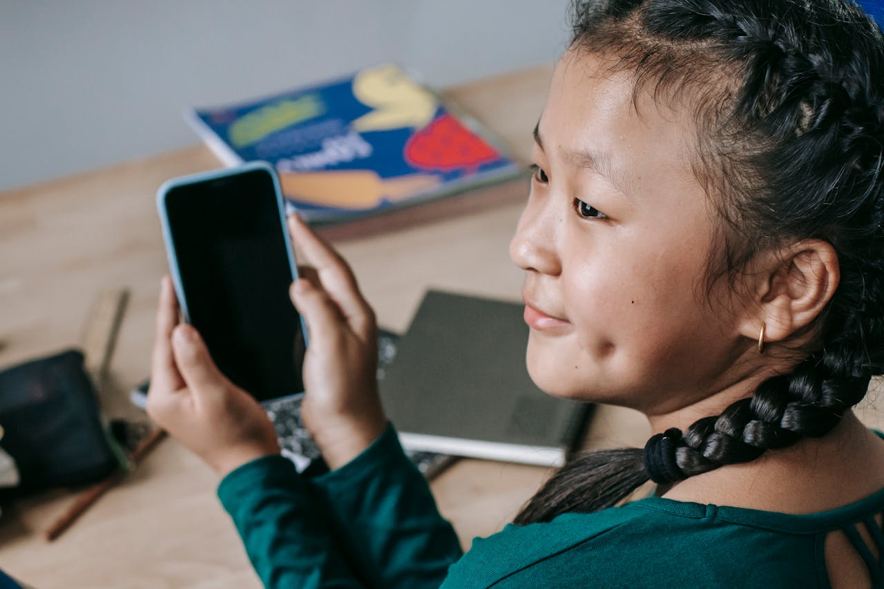 A cheerful girl in braids using a smartphone at her desk, exemplifying modern education and connectivity.