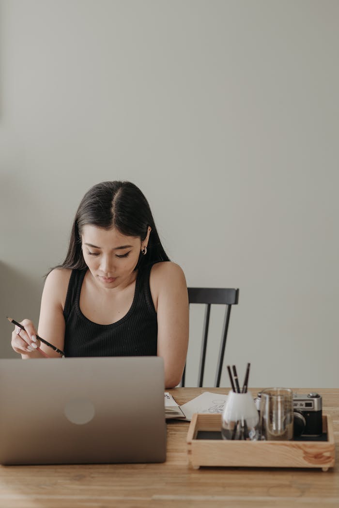Asian woman at home desk engaged in online learning with laptop, notes, and pen.